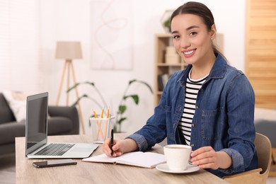 Young woman with cup of drink writing in notebook at wooden table indoors