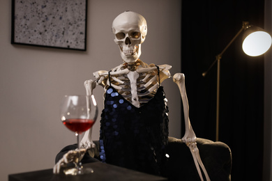 Photo of Skeleton in dress with wine sitting at table indoors