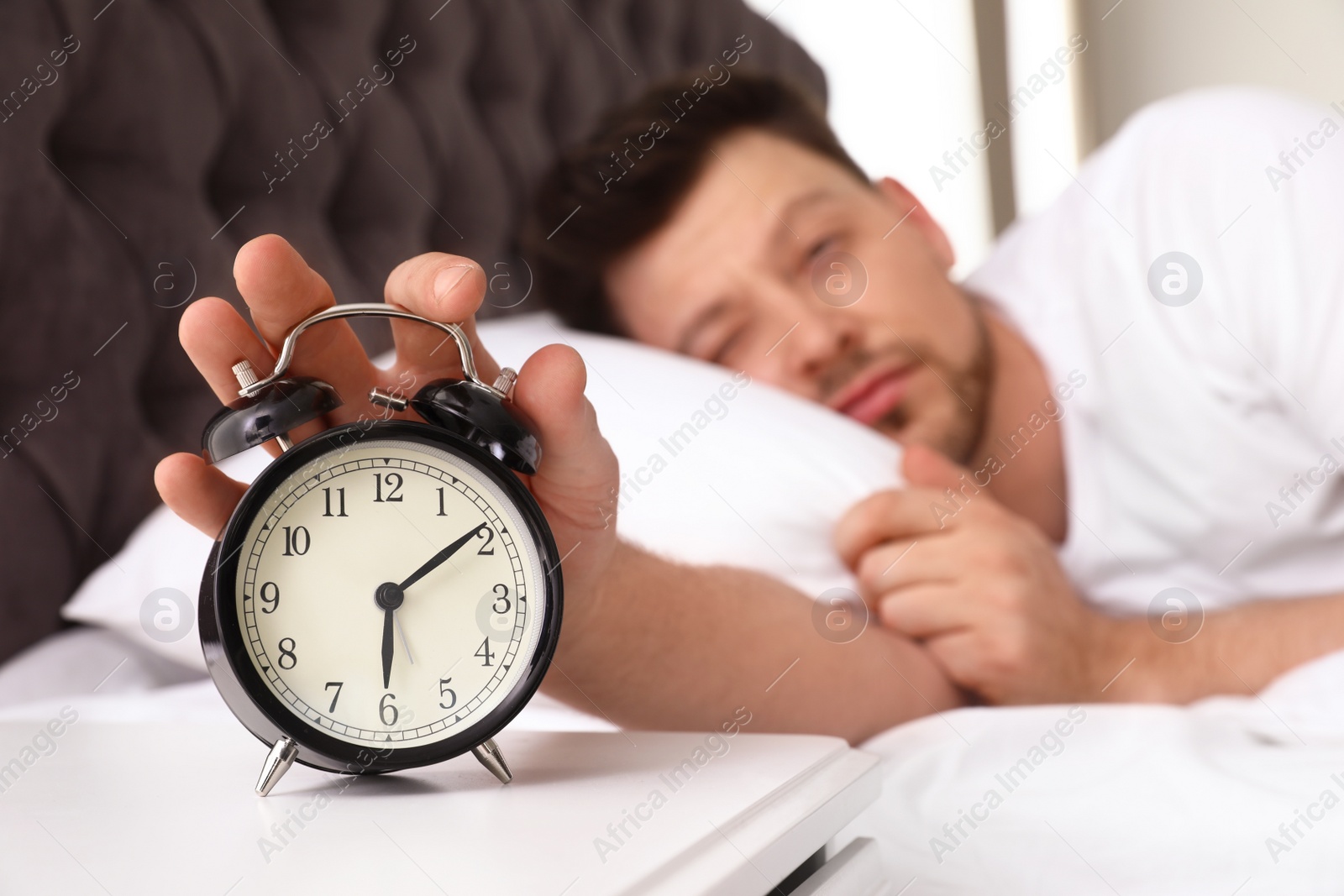 Photo of Sleepy man turning off alarm clock on nightstand. Bedtime