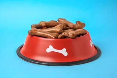 Photo of Red bowl with bone shaped dog cookies on light blue background, closeup
