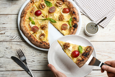 Woman taking slice of delicious cheese pizza with walnuts and grapes at white wooden table, top view