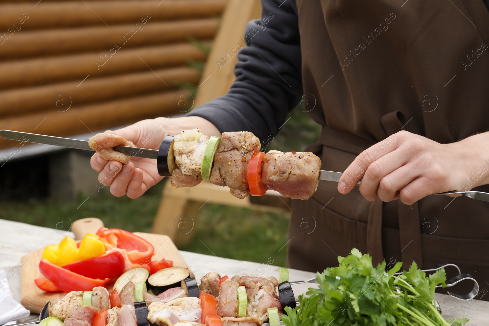 Photo of Woman stringing marinated meat and vegetables on skewer at wooden table outdoors, closeup