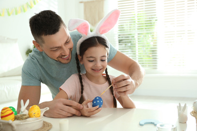 Photo of Happy daughter with bunny ears headband and her father painting Easter egg at home