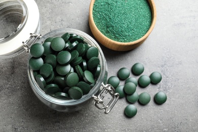 Photo of Flat lay composition of glass jar with green spirulina pills and powder in bowl on grey table