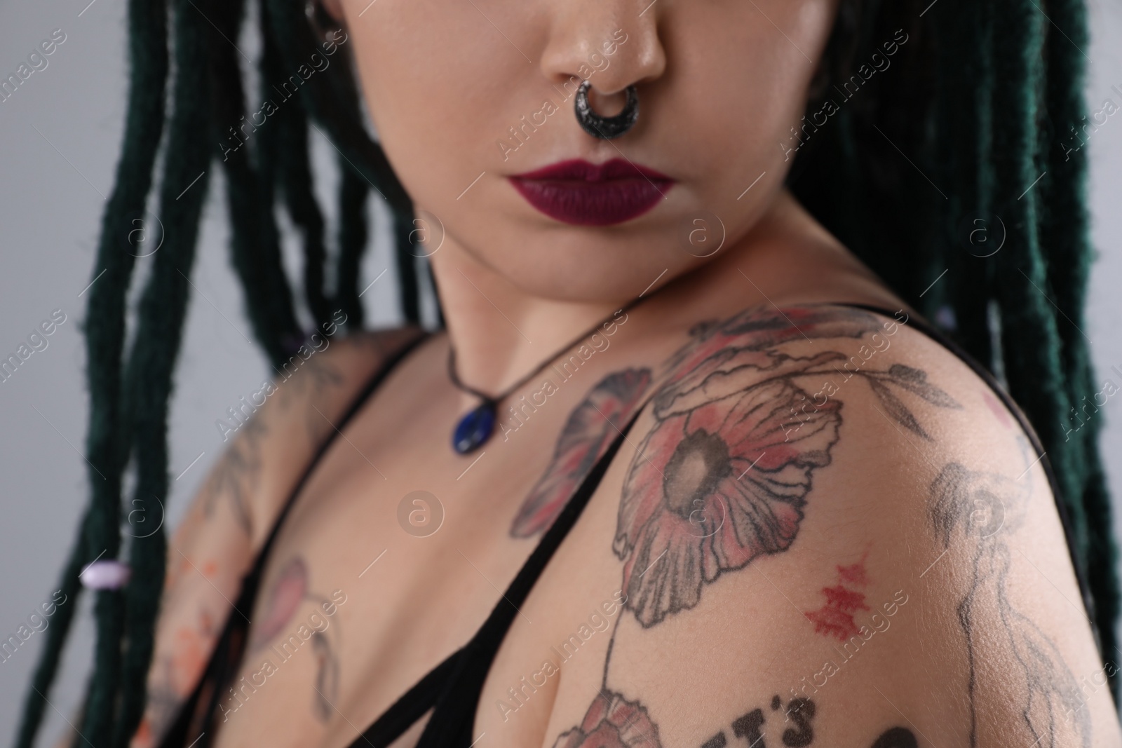 Photo of Young woman with tattoos on body, nose piercing and dreadlocks against grey background, closeup