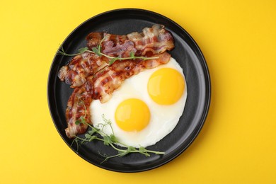 Fried eggs, bacon and microgreens on yellow background, top view