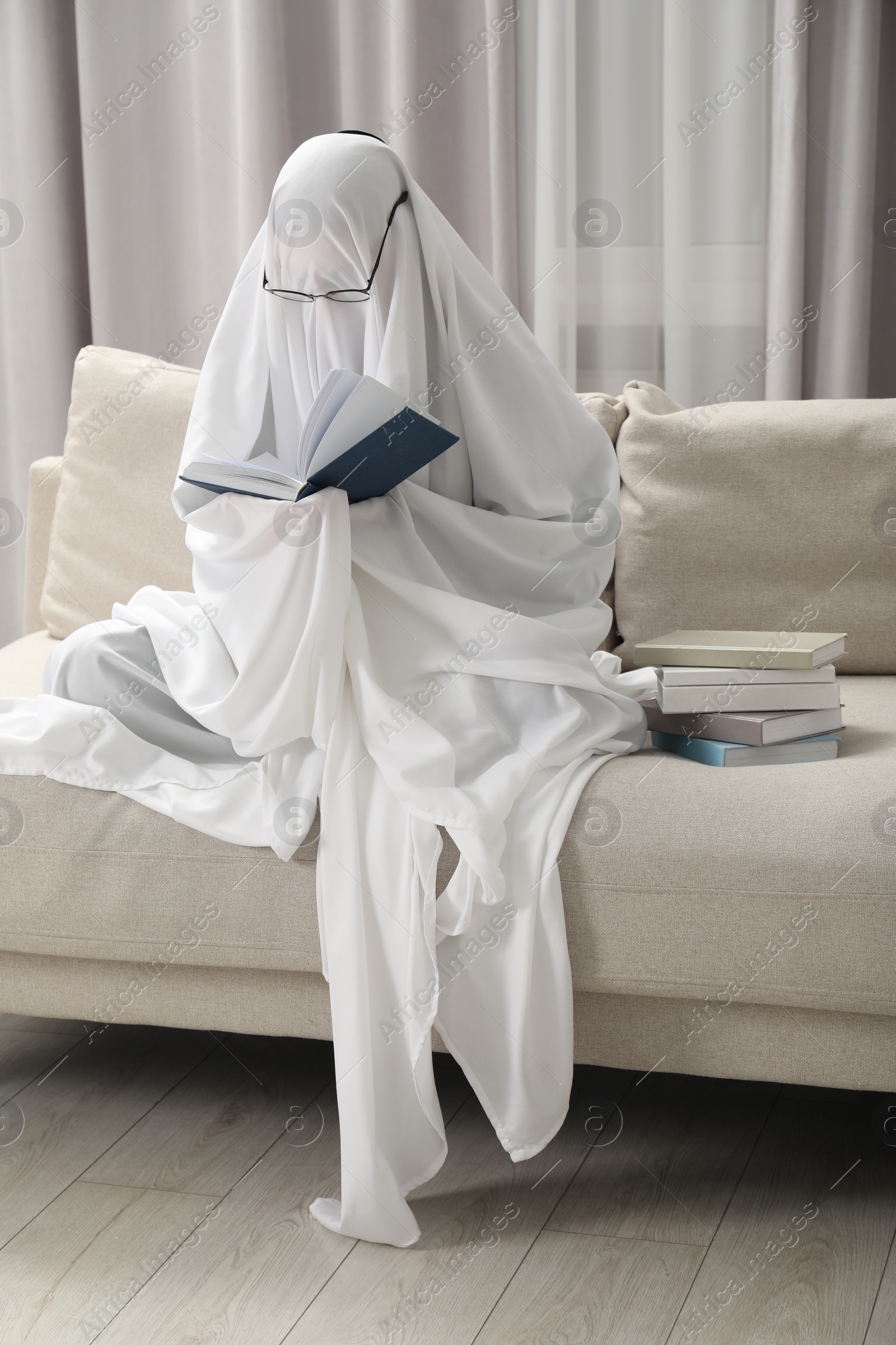 Photo of Creepy ghost. Person covered with white sheet reading book on sofa at home