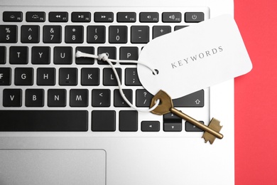 Photo of Laptop, key and tag with word KEYWORDS on red background, top view