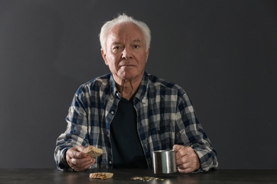 Photo of Poor elderly man with piece of bread and metal mug at table against dark background