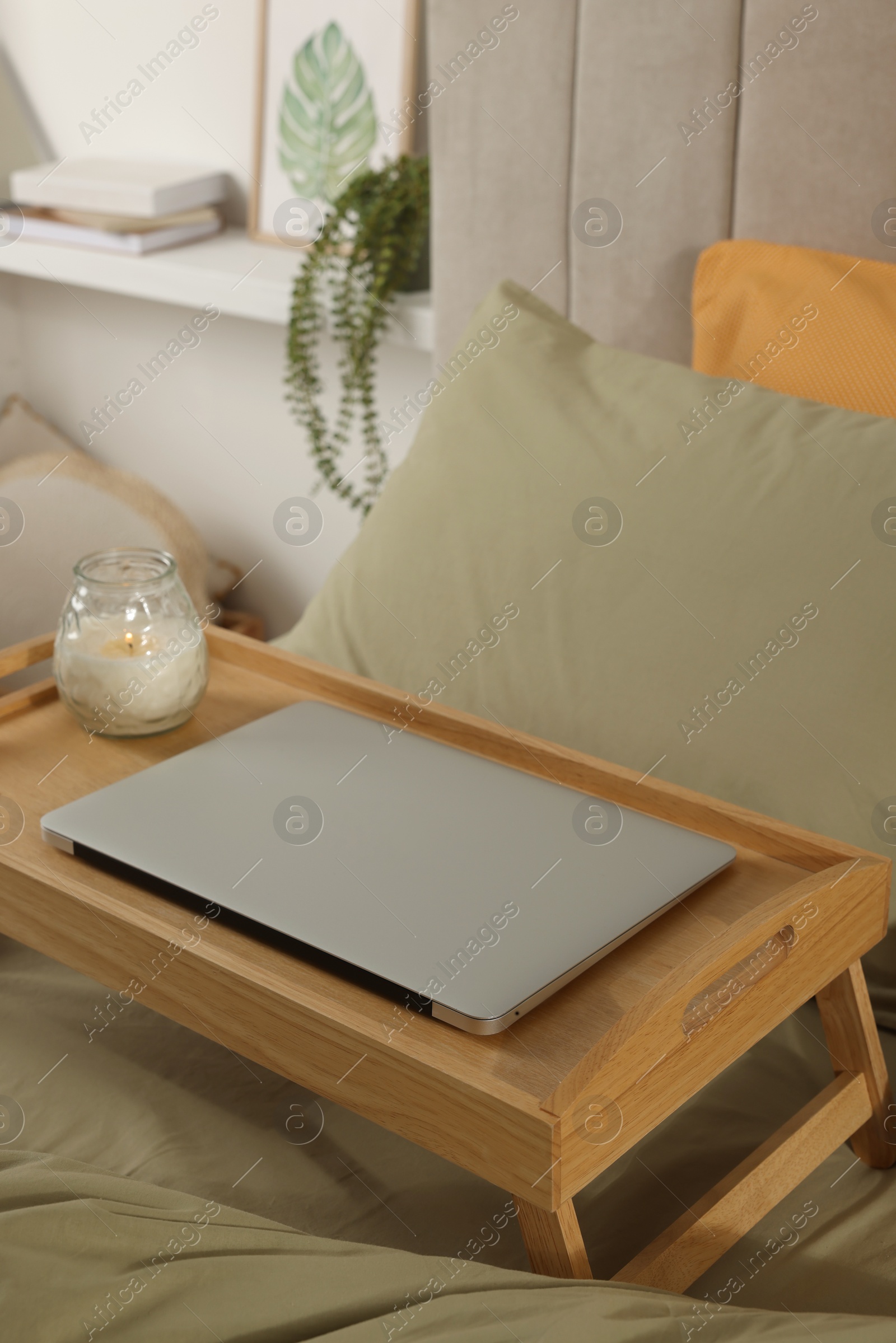 Photo of Wooden tray with modern laptop and burning candle on bed indoors