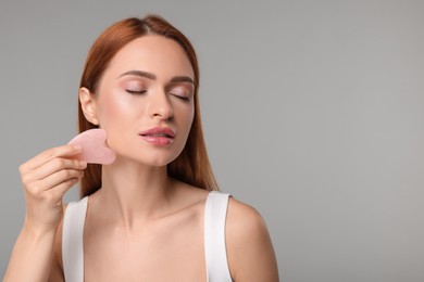 Young woman massaging her face with rose quartz gua sha tool on grey background, space for text