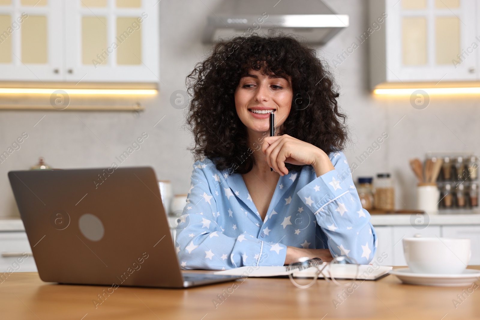 Photo of Beautiful young woman in stylish pyjama with pen using laptop at wooden table in kitchen