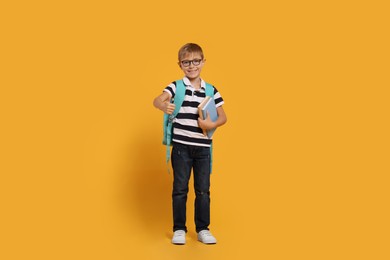 Photo of Happy schoolboy in glasses with backpack and books showing thumb up gesture on orange background