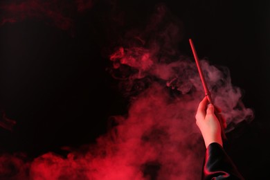 Magician holding wand in smoke on dark background, closeup. Space for text
