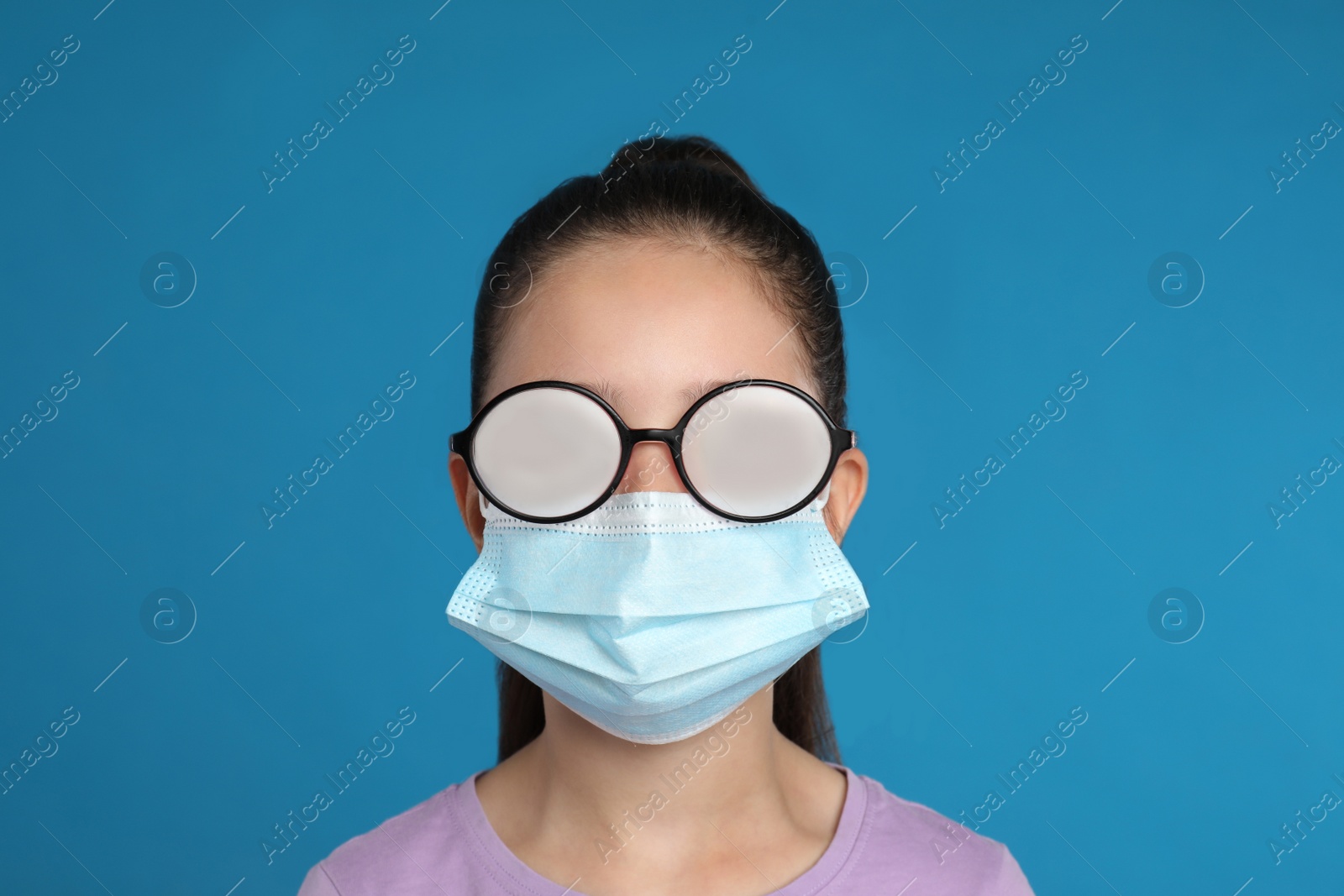 Photo of Little girl with foggy glasses caused by wearing medical face mask on blue background. Protective measure during coronavirus pandemic