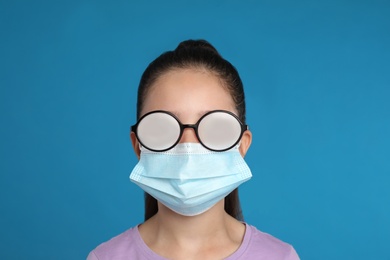 Photo of Little girl with foggy glasses caused by wearing medical face mask on blue background. Protective measure during coronavirus pandemic