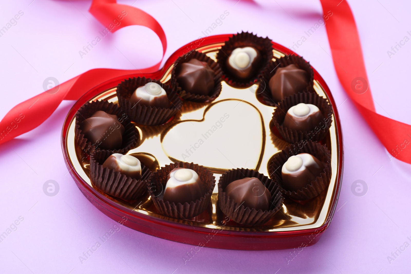 Photo of Heart shaped box with delicious chocolate candies and ribbon on lilac background, closeup