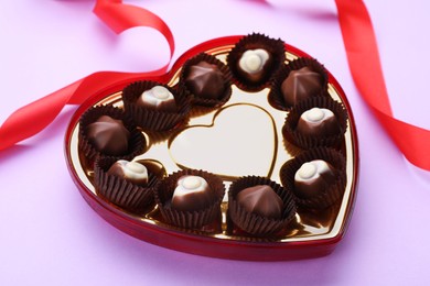 Heart shaped box with delicious chocolate candies and ribbon on lilac background, closeup