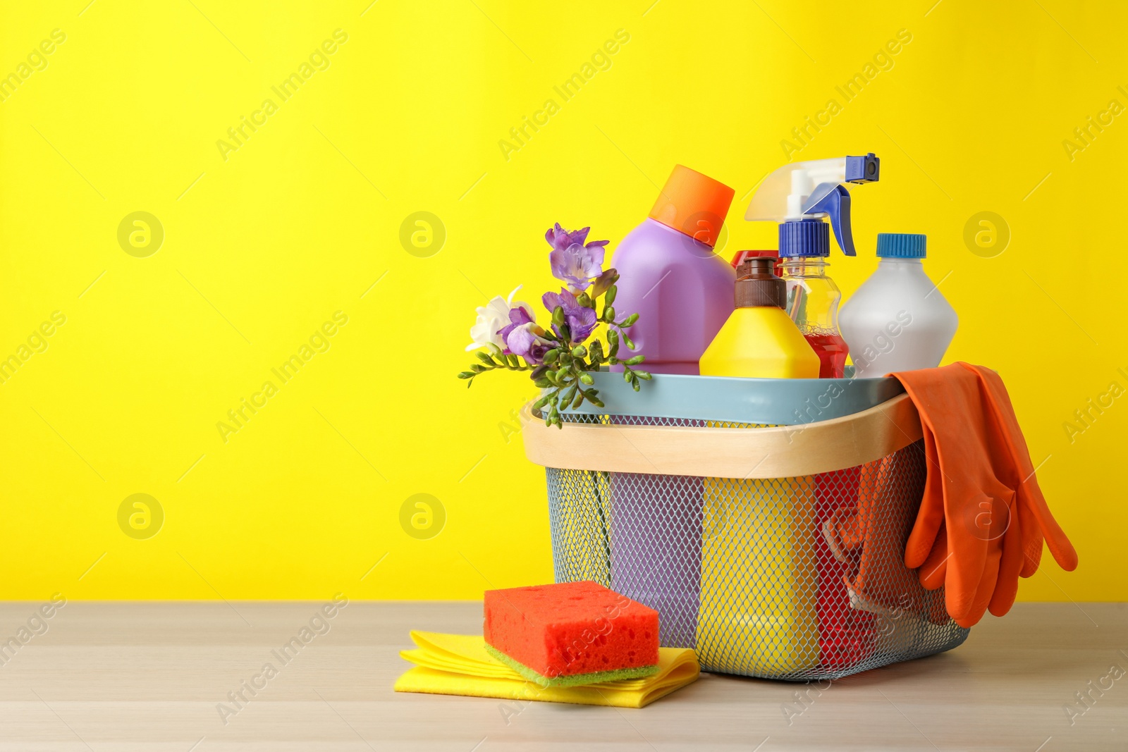 Photo of Plastic basket with spring flowers and cleaning supplies on wooden table. Space for text