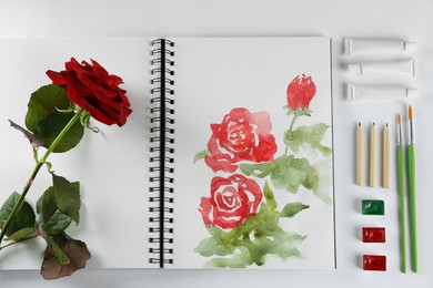 Photo of Painting of roses in sketchbook, flower and art supplies on white background, top view