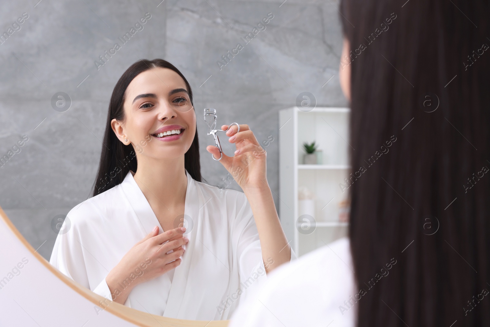 Photo of Beautiful young woman with eyelash curler near mirror in bathroom