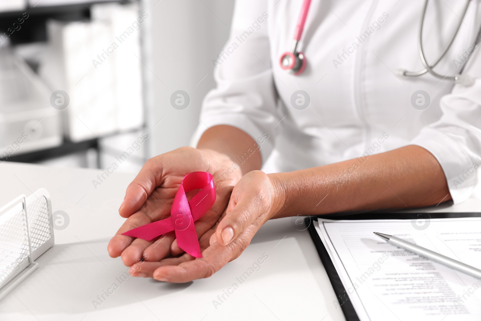 Photo of Doctor holding pink ribbon at white desk indoors, closeup. Breast cancer awareness