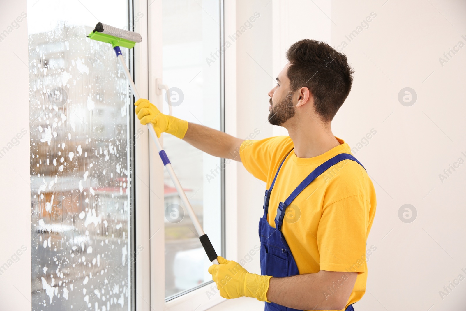 Photo of Professional young janitor in uniform cleaning window indoors