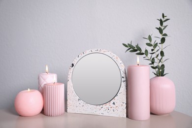 Photo of Small mirror, burning candles and vase on table near light wall
