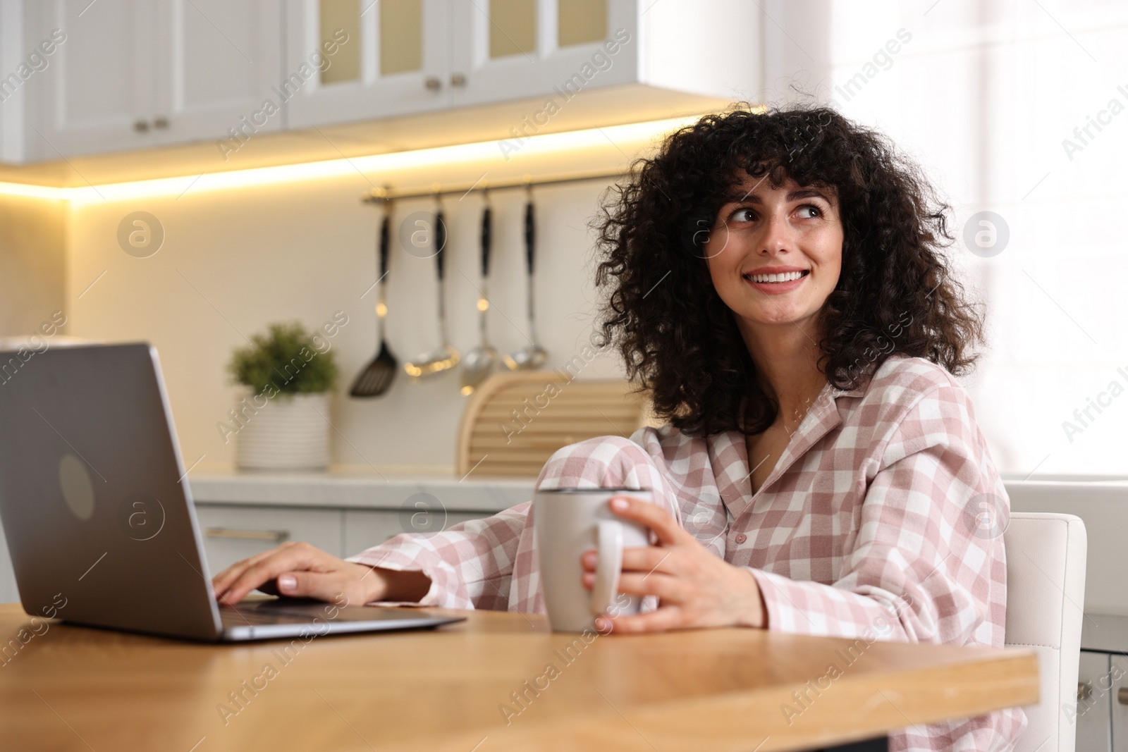 Photo of Beautiful young woman in stylish pyjama with cup of drink using laptop at wooden table in kitchen