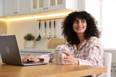 Photo of Beautiful young woman in stylish pyjama with cup of drink using laptop at wooden table in kitchen