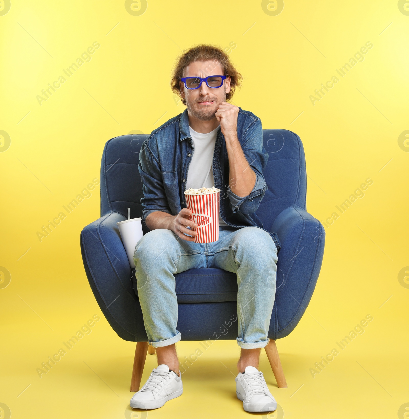 Photo of Emotional man with 3D glasses, popcorn and beverage sitting in armchair during cinema show on color background