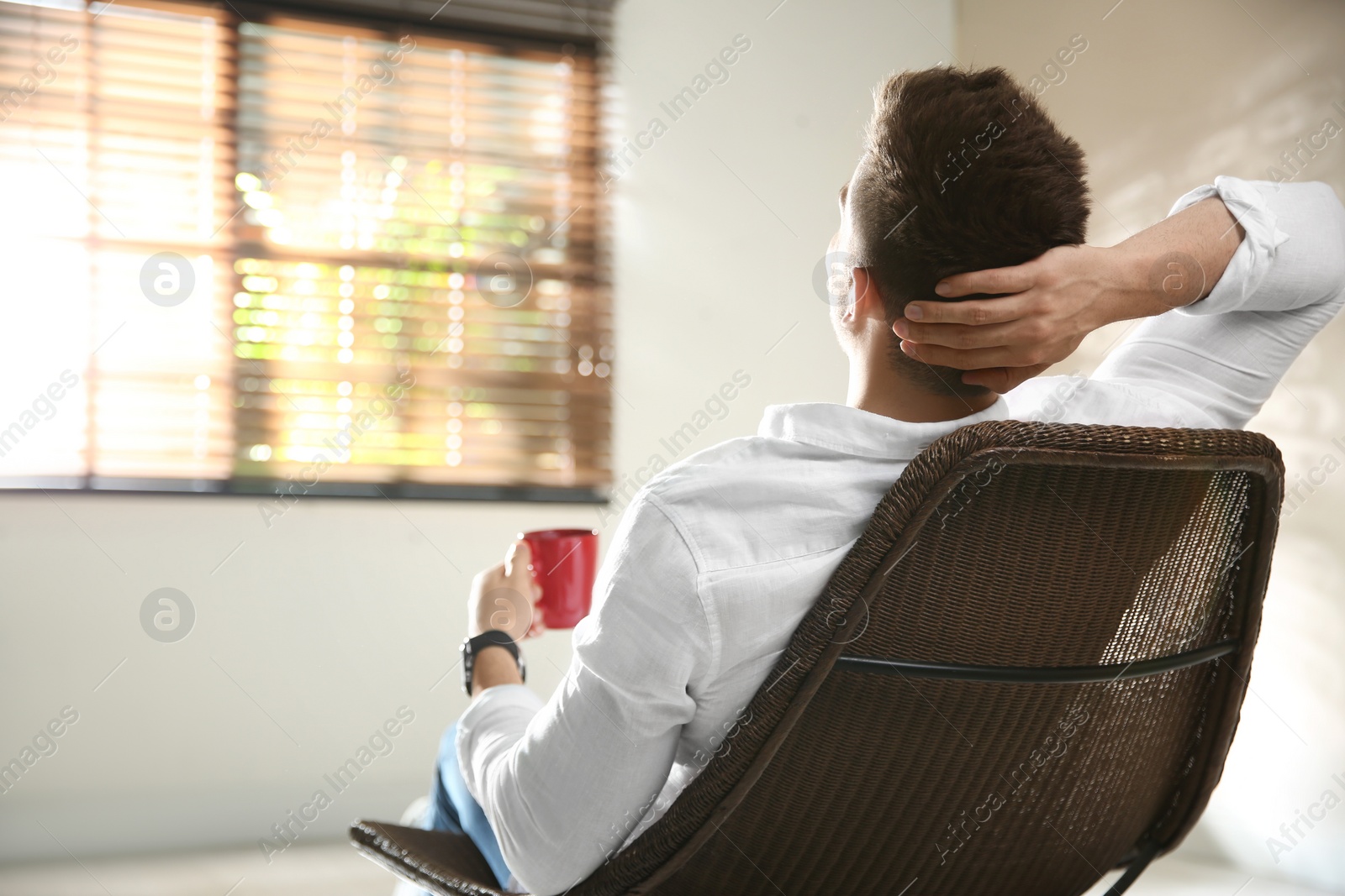 Photo of Young man with cup of drink relaxing near window at home, back view. Space for text