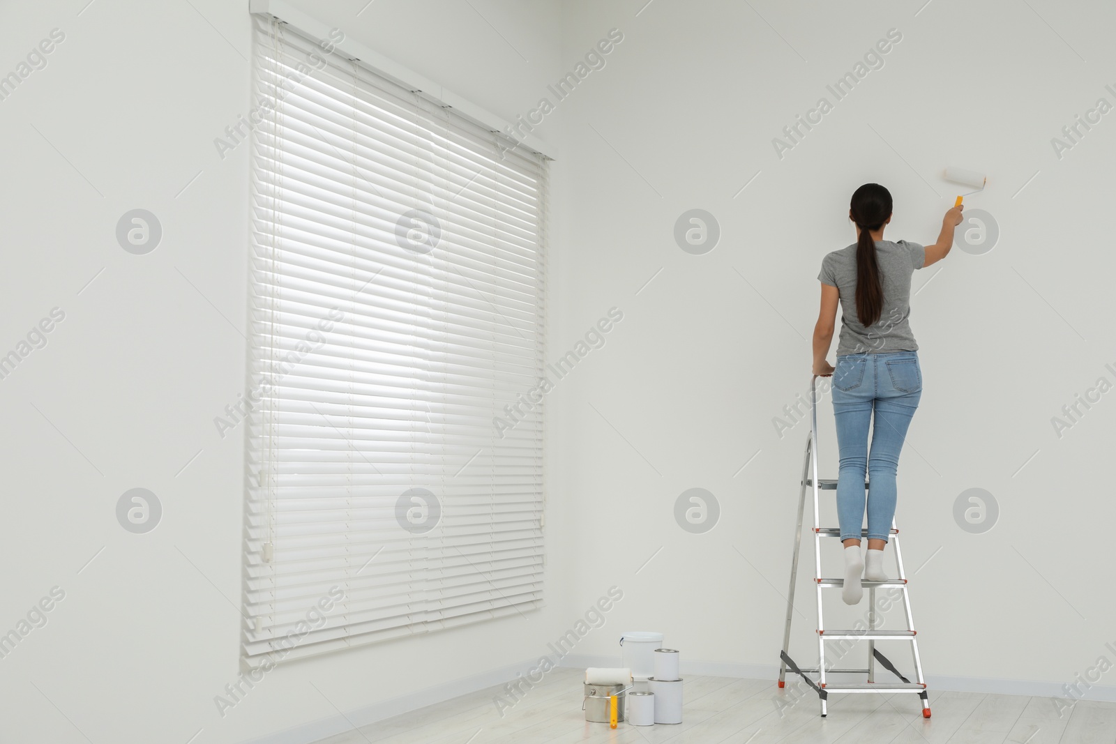 Photo of Woman standing on metallic folding ladder and painting wall indoors, space for text