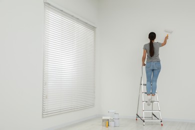 Photo of Woman standing on metallic folding ladder and painting wall indoors, space for text