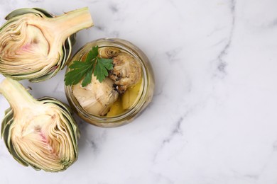 Photo of Jar of delicious pickled artichokes in olive oil and fresh vegetables on white marble table, flat lay. Space for text