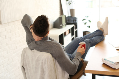 Young man with cup of drink relaxing at table in office during break