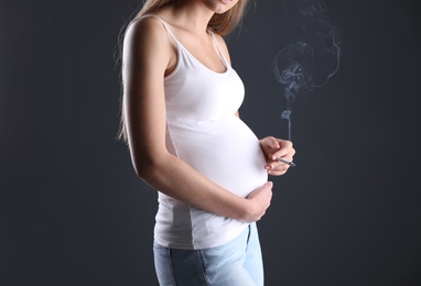 Photo of Young pregnant woman smoking cigarette on dark background, closeup. Harm to unborn baby