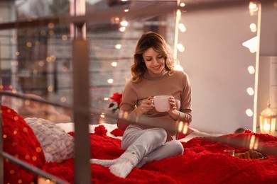Photo of Beautiful young woman with cup of drink relaxing on bed at home. Cozy atmosphere