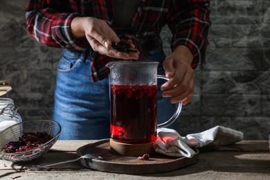 Photo of Woman putting dry flower petals into teapot with hibiscus tea at wooden table, closeup