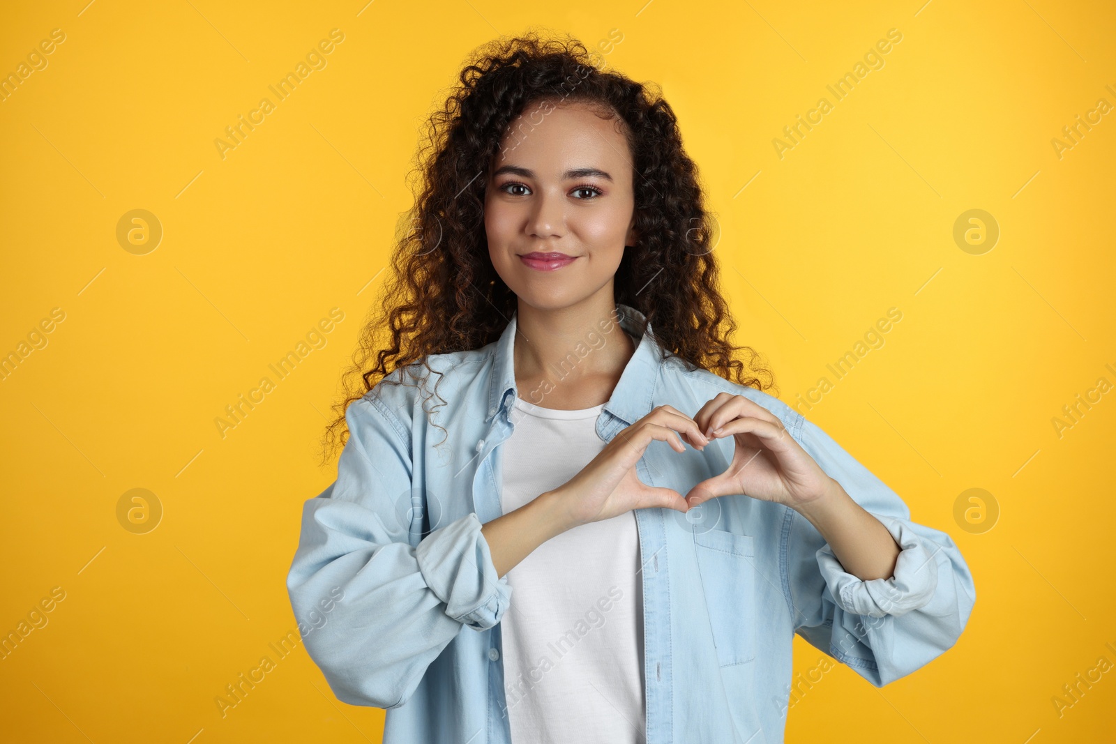Photo of Happy young African-American woman making heart with hands on yellow background