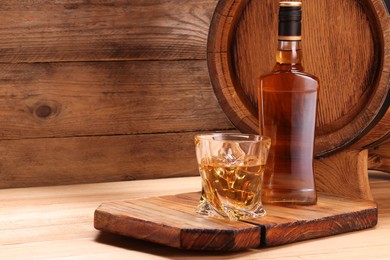 Photo of Whiskey with ice cubes in glass, bottle and barrel on wooden table, space for text