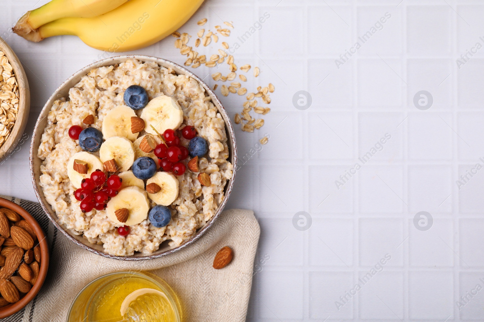 Photo of Bowl of oatmeal berries, almonds and banana slices on white tiled table, flat lay. Space for text