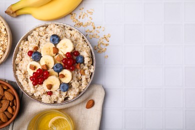 Bowl of oatmeal berries, almonds and banana slices on white tiled table, flat lay. Space for text