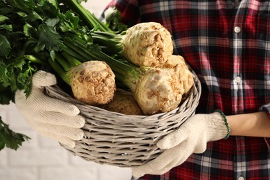 Photo of Woman holding wicker basket with raw celery roots near white brick wall, closeup
