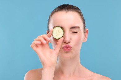 Photo of Beautiful woman covering eye with piece of cucumber and blowing kiss on light blue background