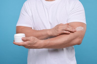 Man applying body cream onto his arm on light blue background, closeup