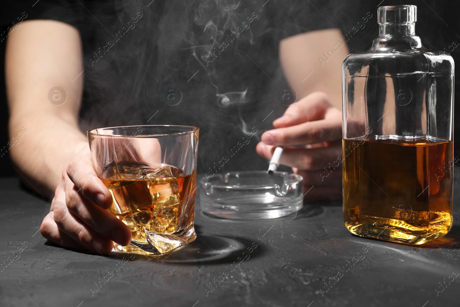 Photo of Alcohol addiction. Man with whiskey and smoldering cigarette at dark textured table, closeup