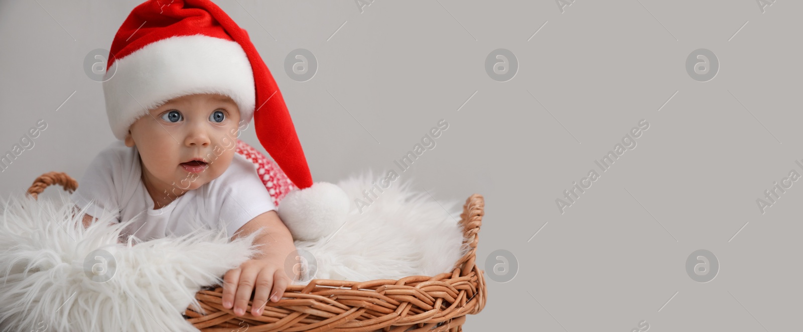 Image of Cute baby wearing Santa hat in wicker basket on light grey background, banner design with space for text. Christmas celebration