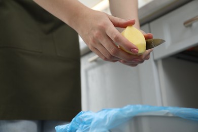 Woman peeling fresh potato with knife above garbage bin indoors, closeup