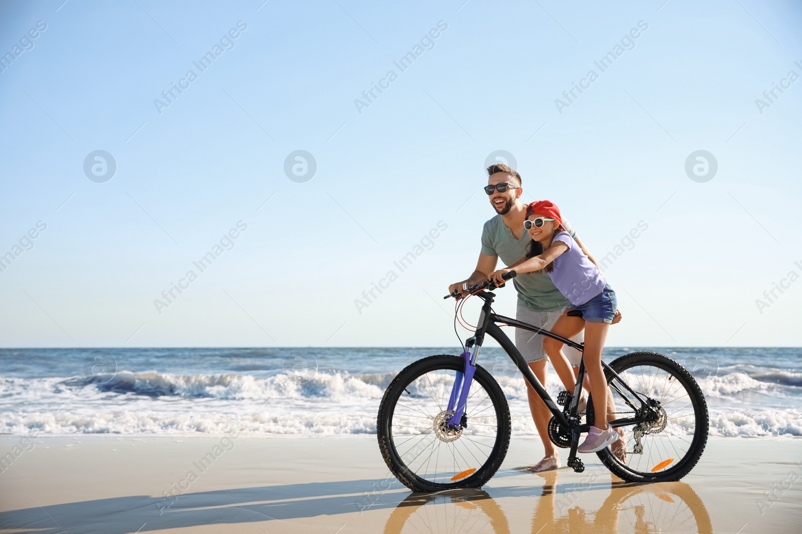 Photo of Happy father teaching daughter to ride bicycle on sandy beach near sea